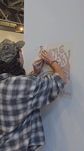 Person wearing a camouflage hat and plaid shirt writing or drawing on a poster on a white wall.