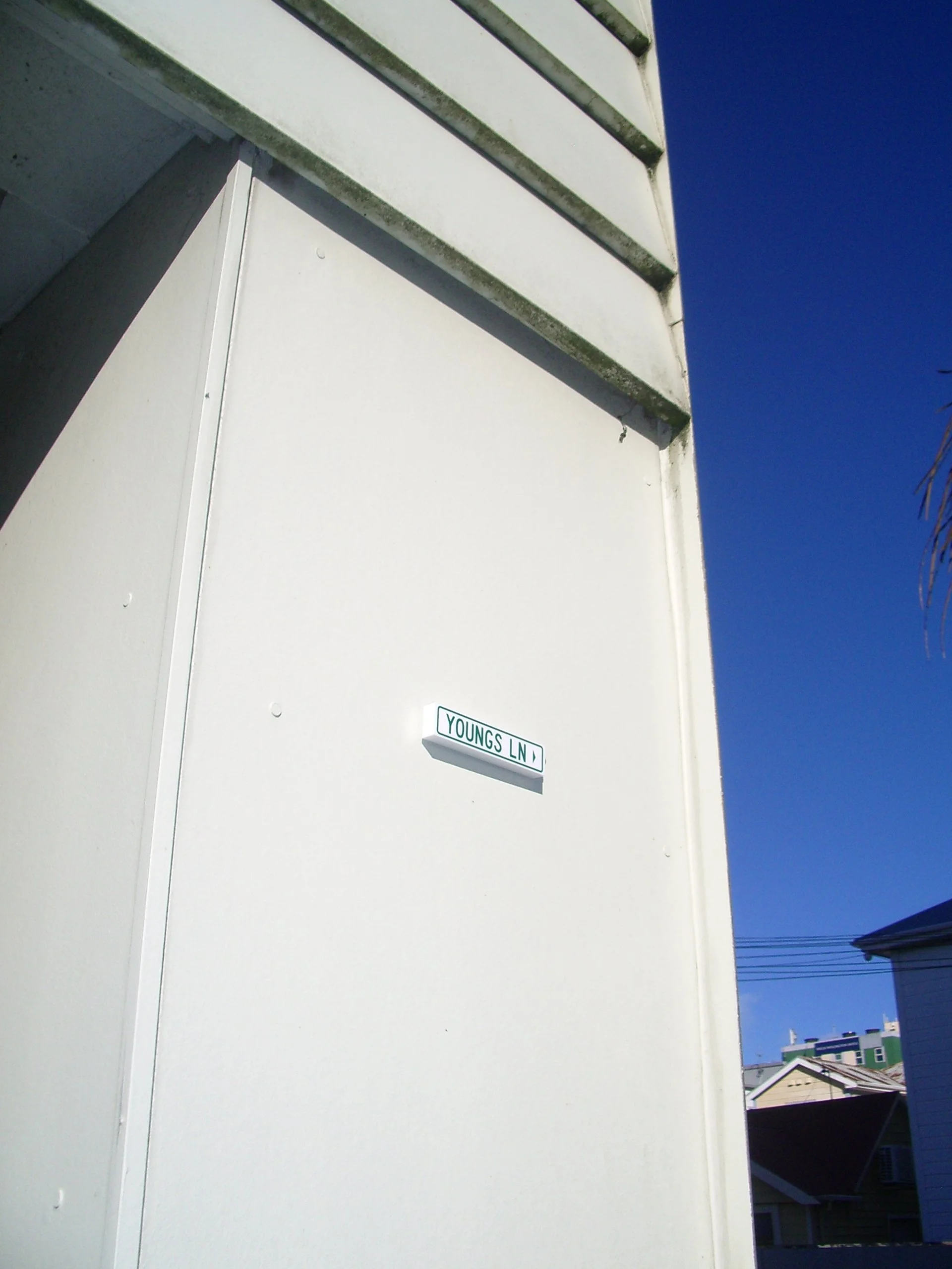 A street sign reading 'Youngs Ln' mounted on a white exterior wall of a building, with blue sky and rooftops visible in the background.