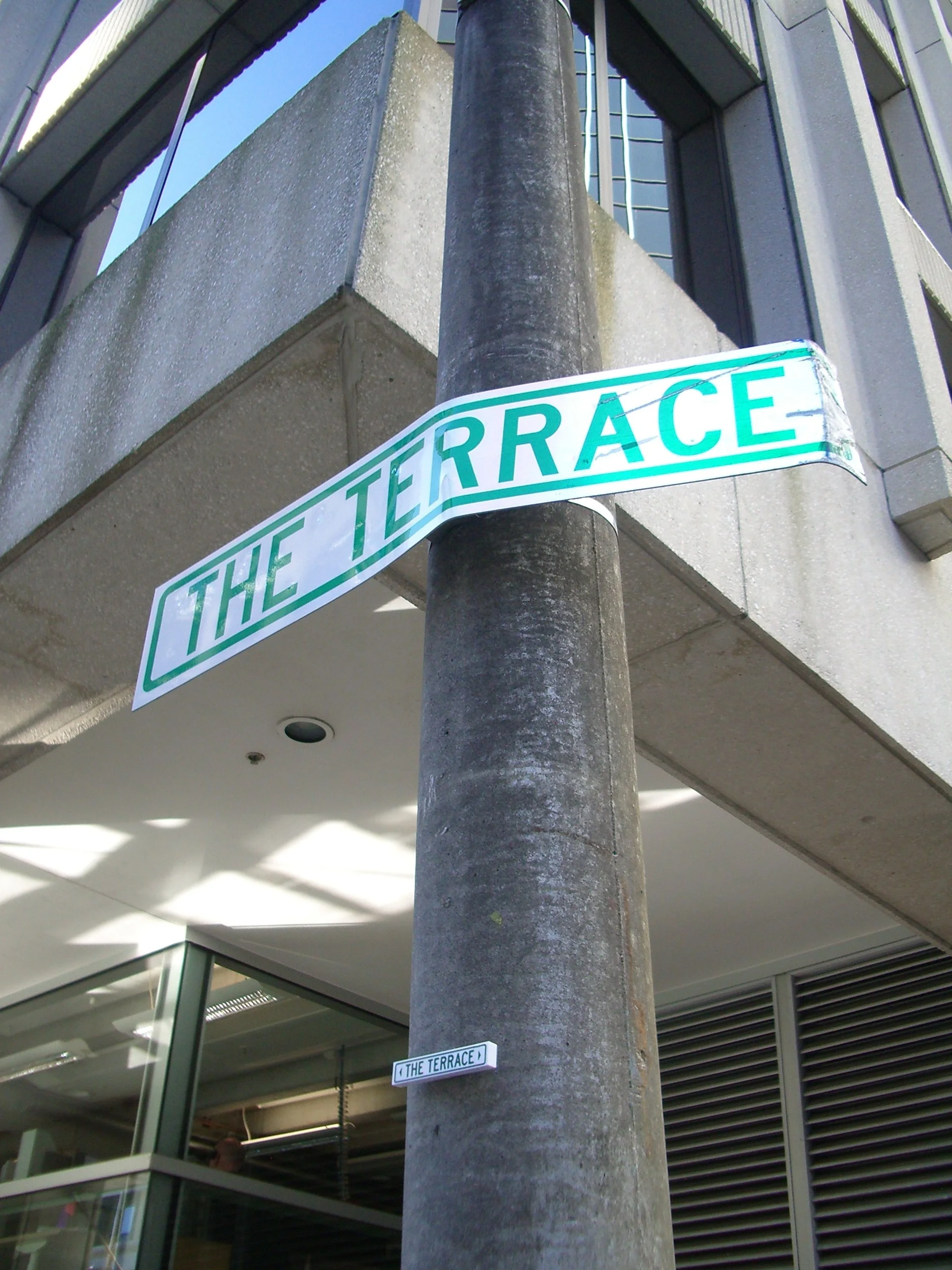 Street corner with a traffic pole and street signs that read 'THE TERRACE.' Modern buildings with glass windows and metallic panels surround the corner.