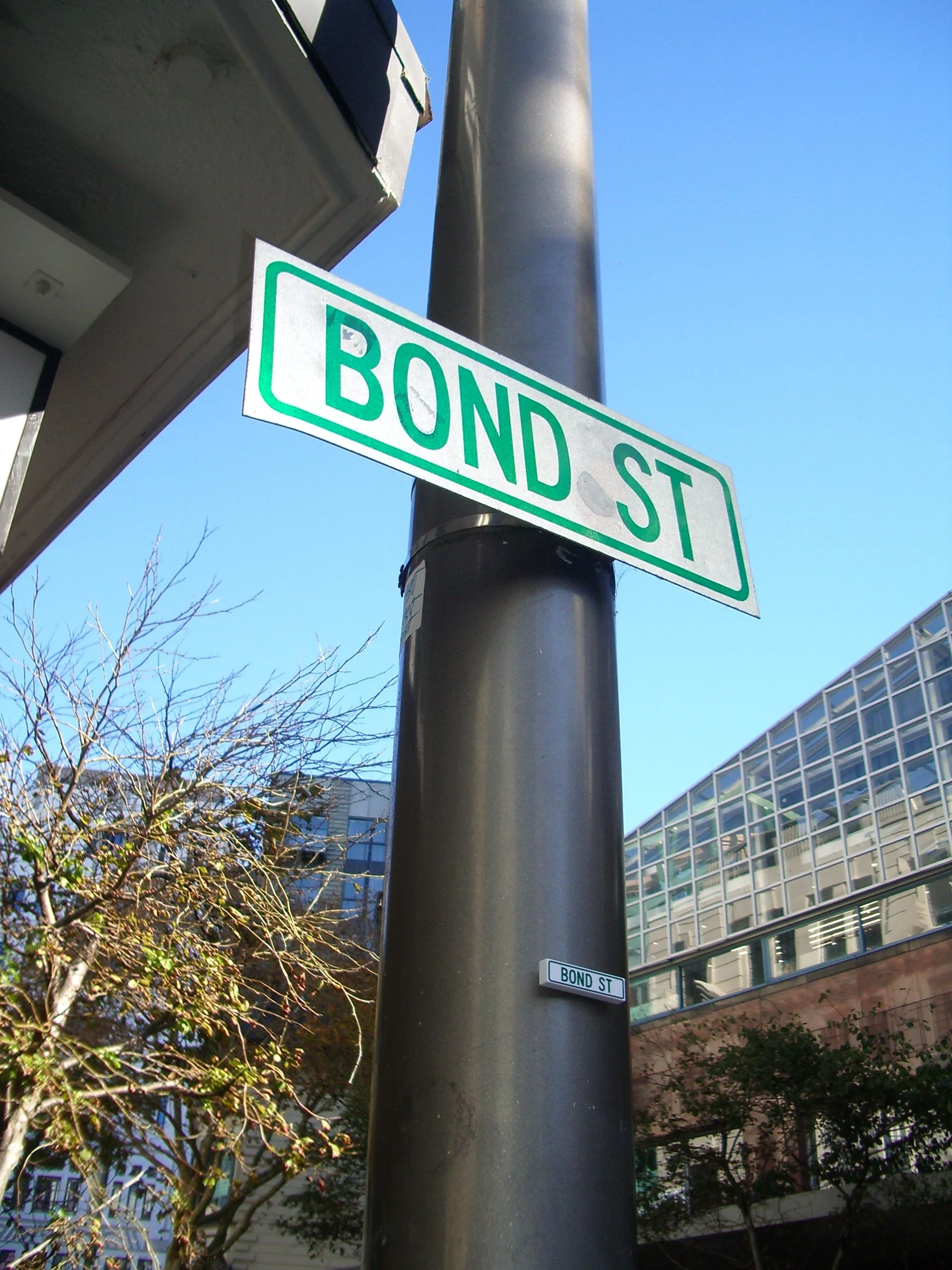 Street sign on a pole at the intersection of Bond Street, with a modern building with glass windows and trees in the background under a clear blue sky.