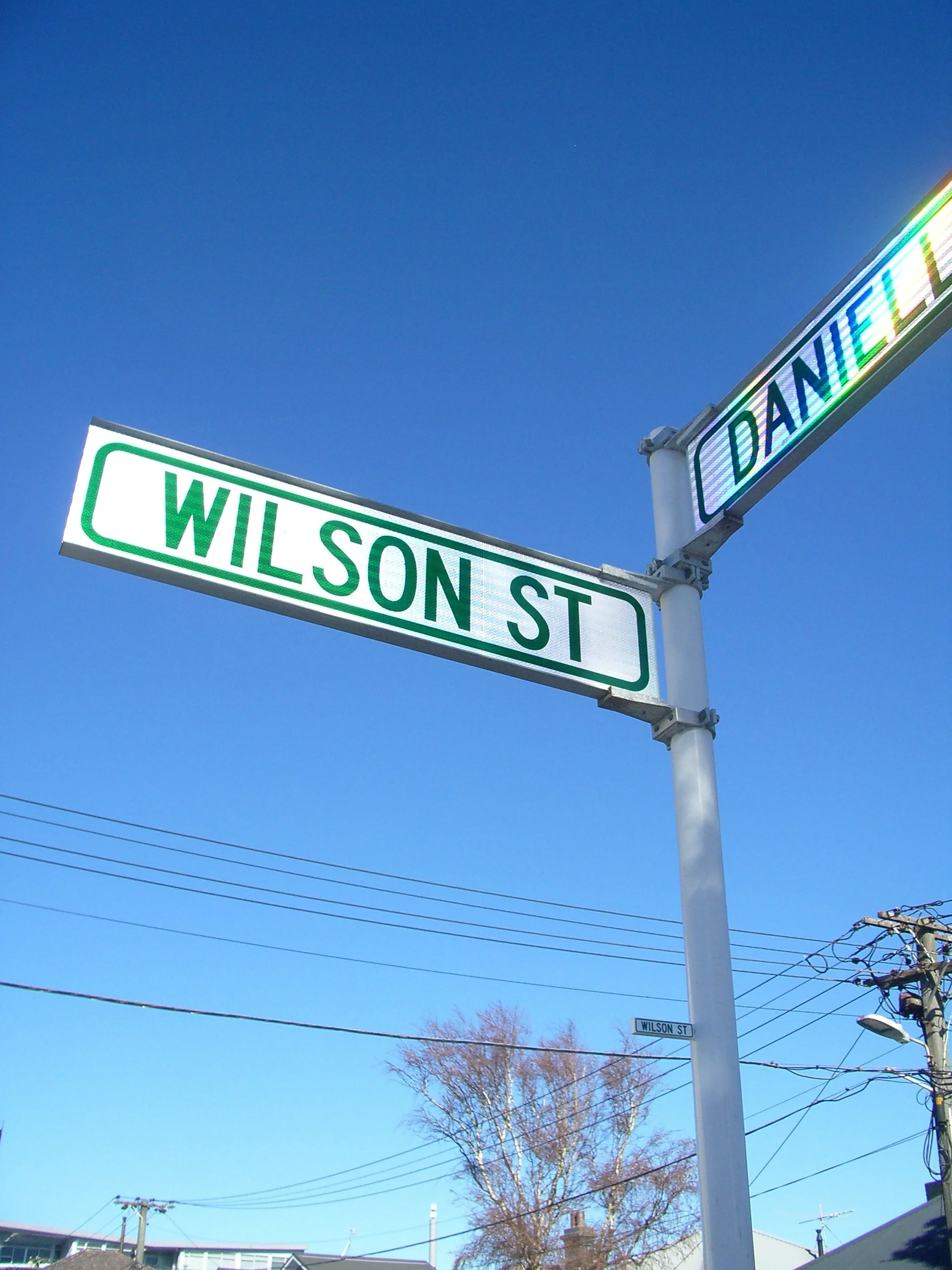 Street signs at the intersection of Wilson Street and Daniel Street against a bright blue sky. There are utility poles and power lines, with a tree and buildings visible in the background.