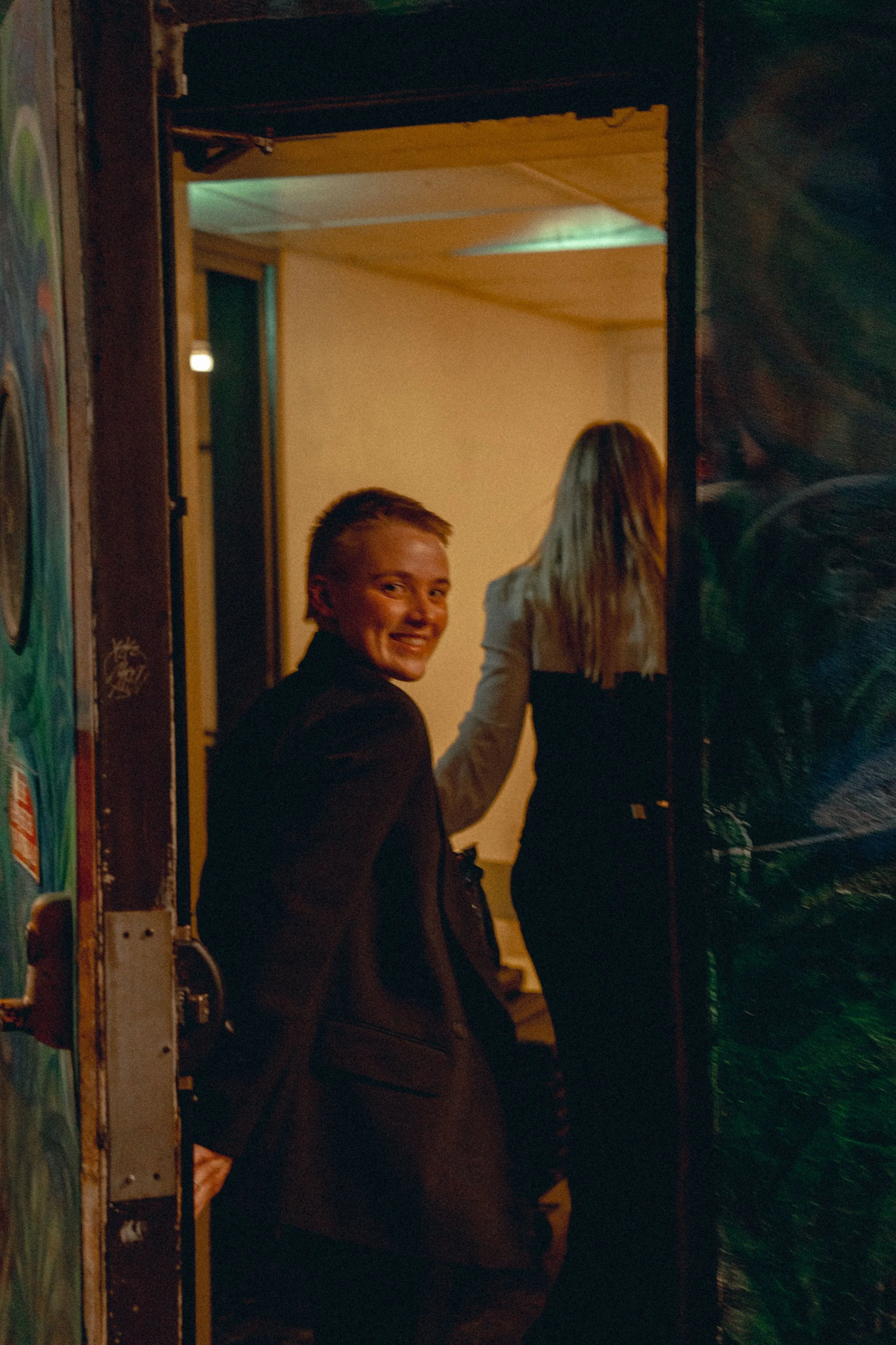 A young woman with short blond hair smiling, and a woman with long blond hair, both standing at a doorway in a backstage. The young woman is wearing a dark jacket, and the woman is wearing a white top. They are members of the band CHARLOT.