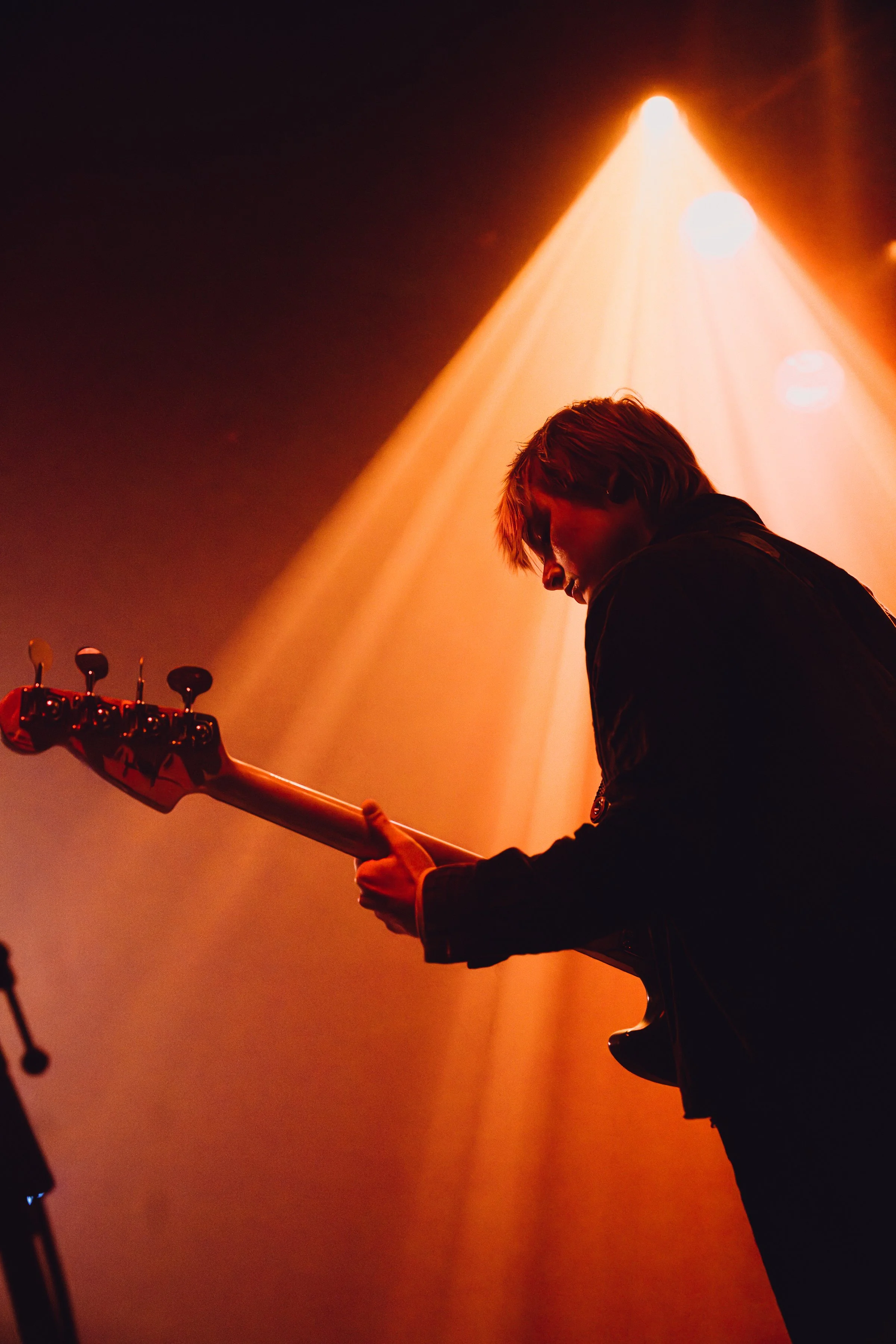 A musician playing an electric bass guitar on stage under orange stage lights, with focus on the instrument and the person's silhouette, with the band Cocobolo.
