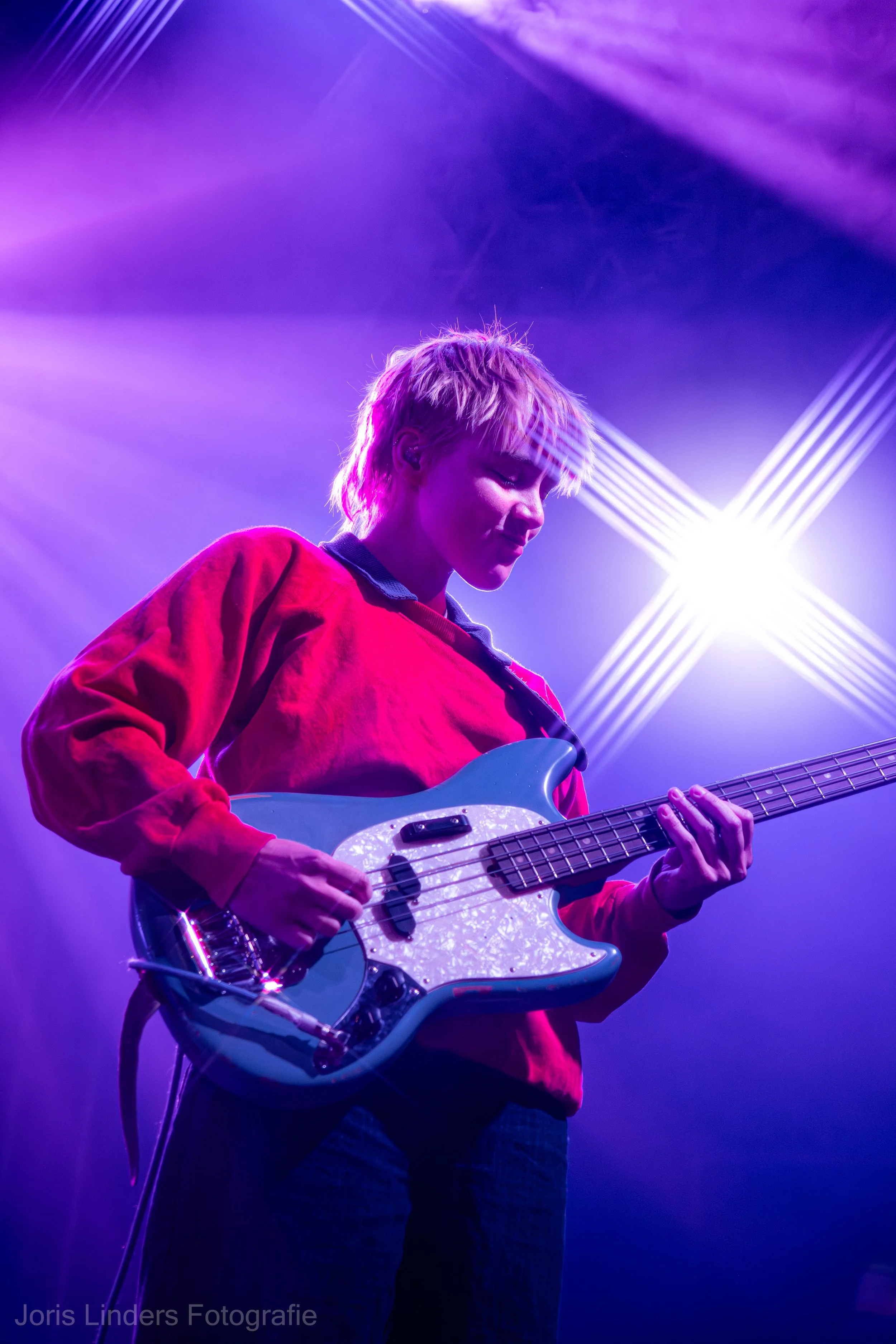 A person with blonde hair playing an electric bass guitar on stage with purple and blue lighting and beams of light in the background with a Mustang bass guitar.