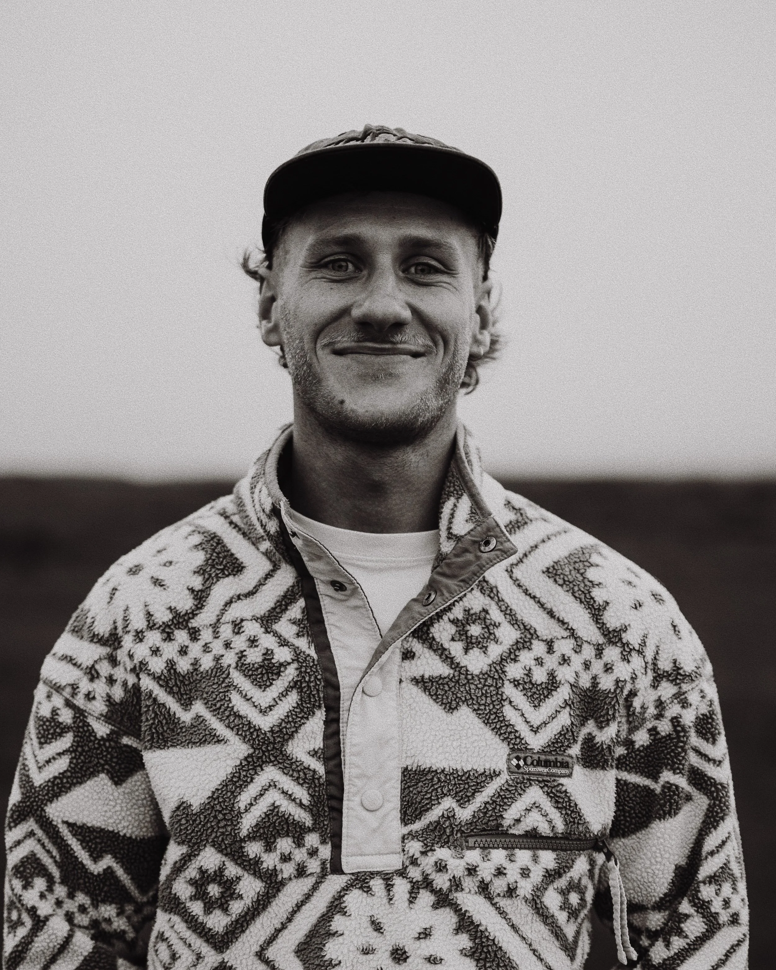 Homme souriant portant une casquette et un pull en laine avec un motif géométrique, debout en extérieur.