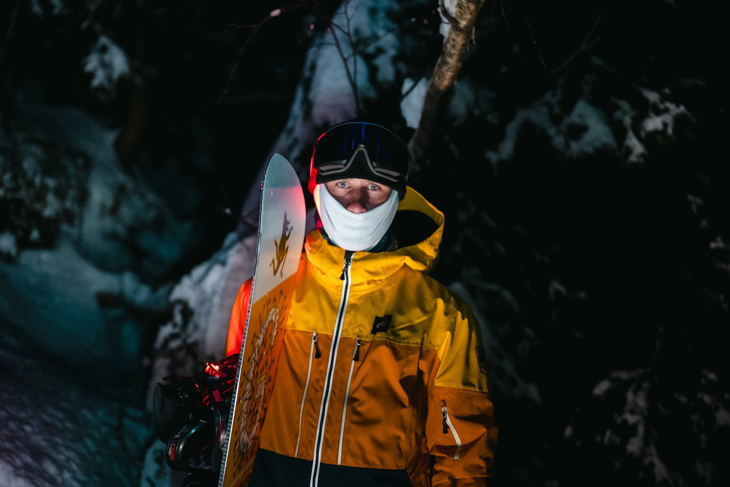 Un skieur en tenue jaune, portant un casque et un masque, se tient dans la neige sombre, regardant vers l'appareil photo dans une forêt enneigée lors de la nuit.