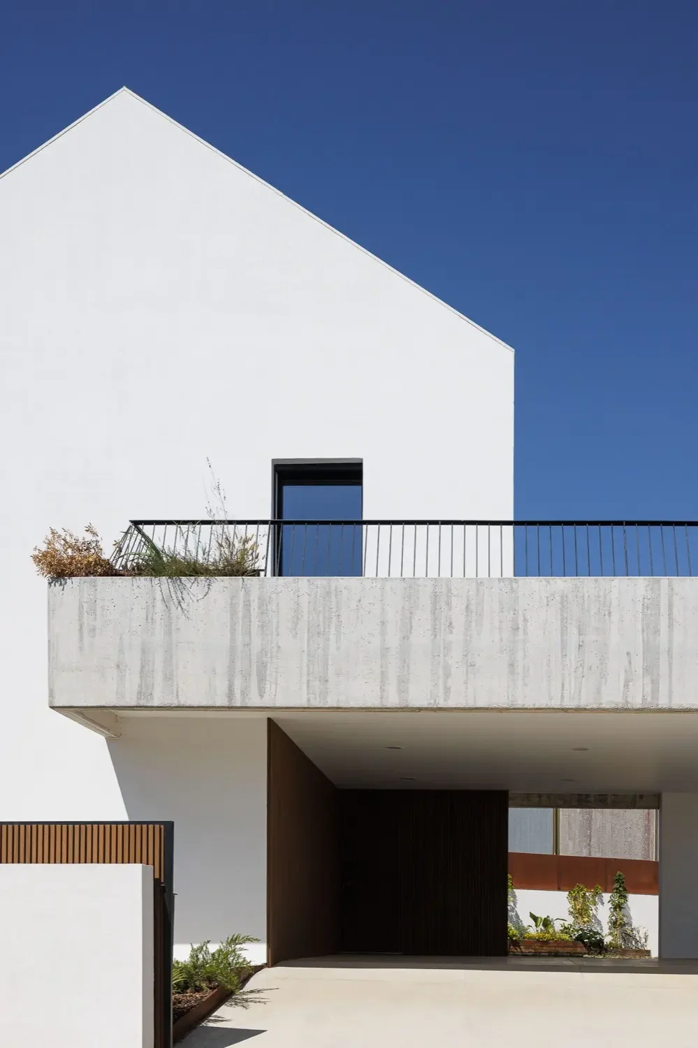 Modern white building with a sloped roof, a small balcony with black railing, and plants on the balcony. Clear blue sky in the background.