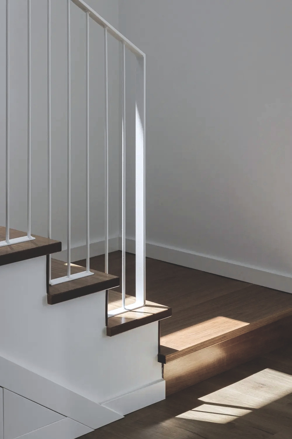 Part of a staircase with dark wood steps, white risers, and a white metal railing, beside a corner with a gray wall and wooden flooring.
