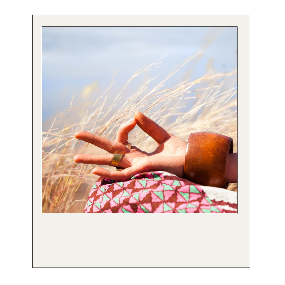 Close-up of a hand with a ring and a large bracelet, making a gesture with three fingers, in a field of tall, dry grass under a clear sky.