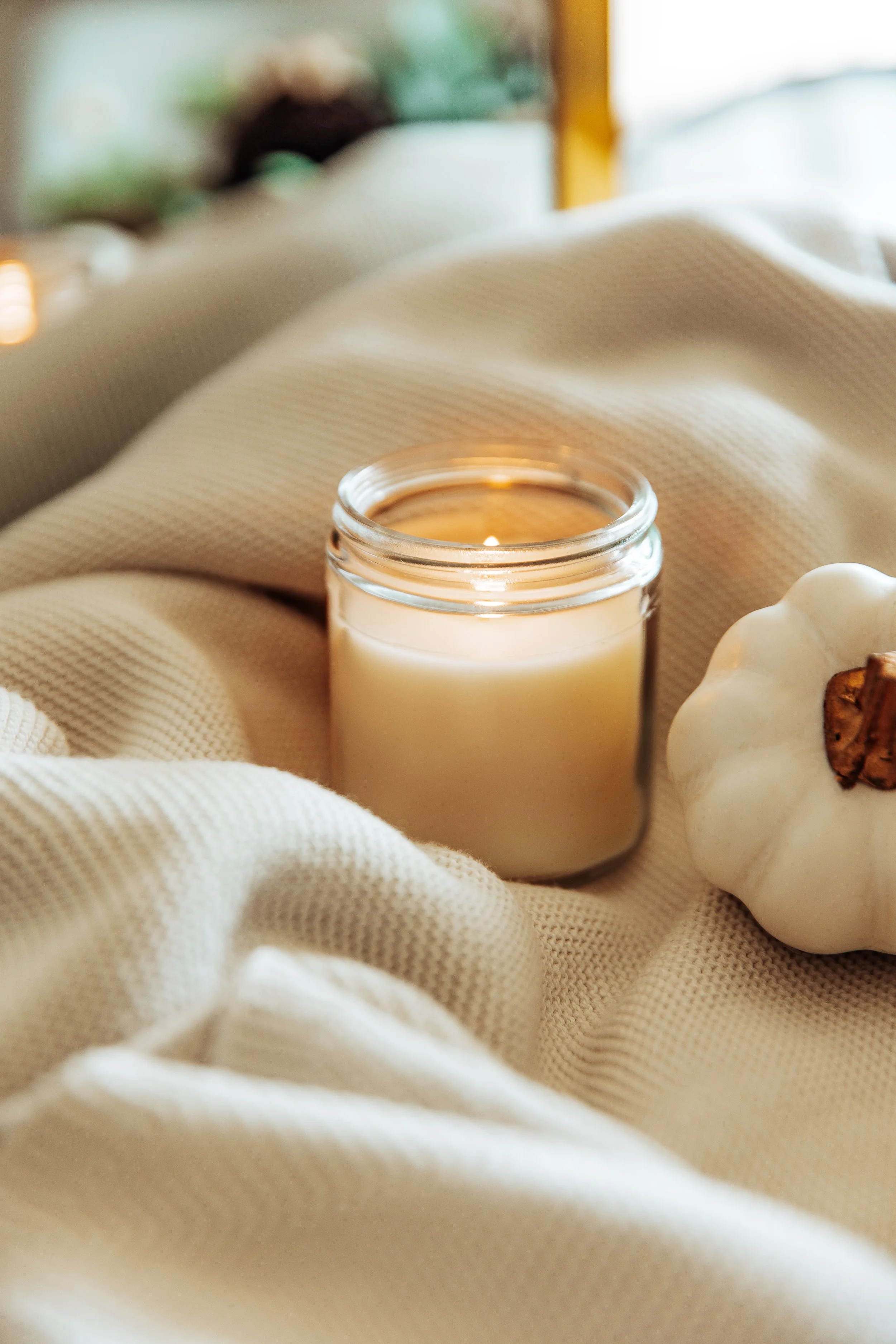 Close-up of a lit beige candle in a glass jar and a white pumpkin with a brown stem, resting on a textured beige fabric.