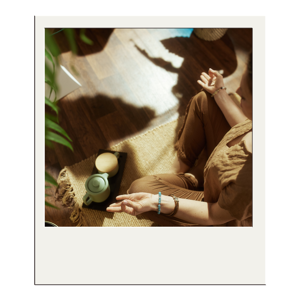 A woman sitting cross-legged on a woven rug, practicing meditation with her hands resting on her knees, palms facing upward. She is wearing a brown shirt and brown pants, with bracelets on her wrists. On the rug beside her, there is a small black tray with a green teapot and a light-colored container. There is a potted plant in the foreground on the left side, and sunlight is casting shadows on the dark wooden floor.