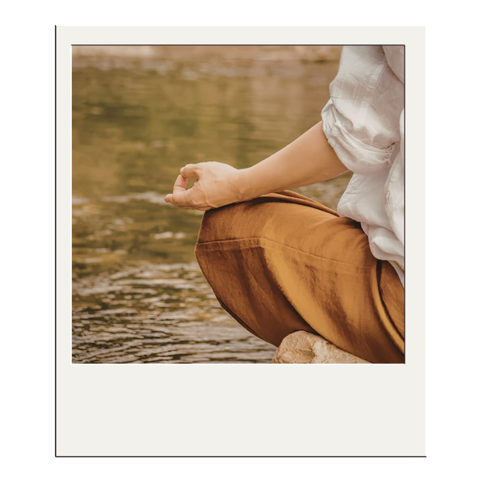 Person practicing meditation by a river, sitting cross-legged with hand resting on knee, wearing a white shirt and tan pants.
