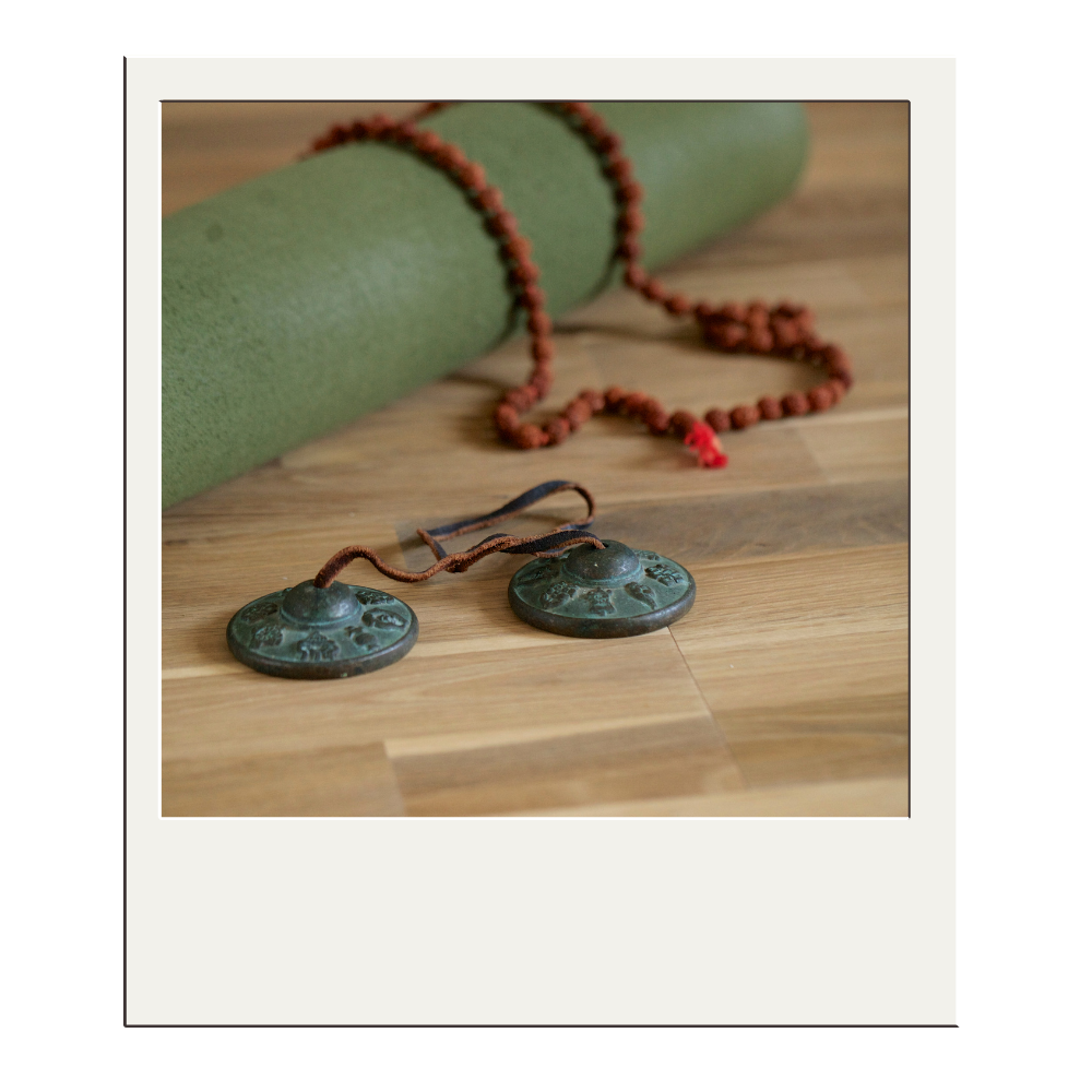 A set of Tibetan tingsha cymbals with a mala prayer bead necklace placed on a wooden surface. A green yoga mat is rolled up in the background.