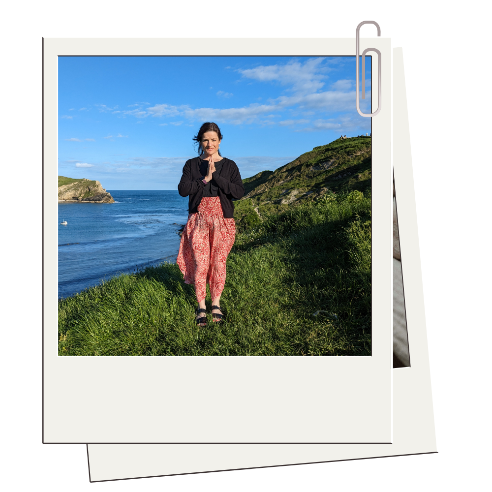 Woman standing on grassy hill near coast with blue sky and ocean in background, hands in prayer position, wearing black top, red patterned pants, and sandals.