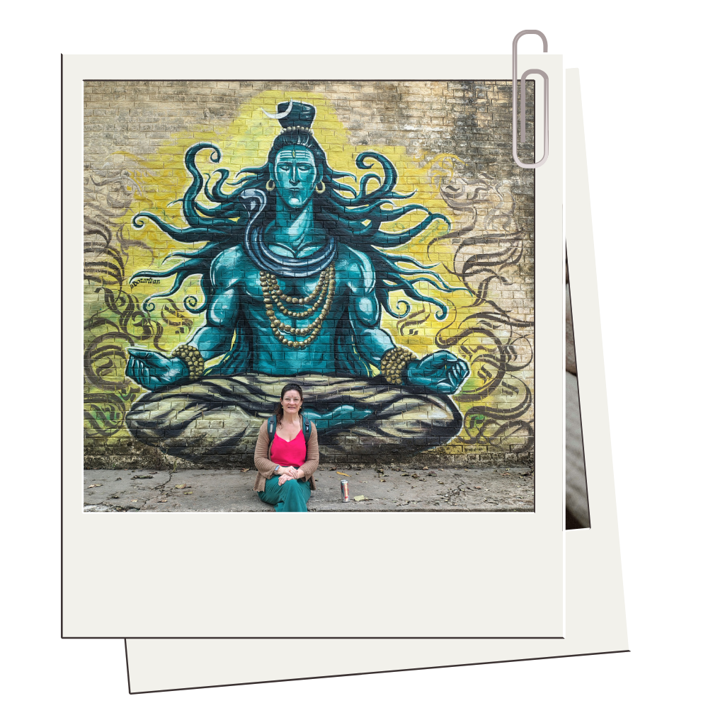 A woman with dark hair, wearing a pink top, brown cardigan, and teal pants, sitting in front of a colorful mural of a meditative blue deity with multiple snakes, on a brick wall.