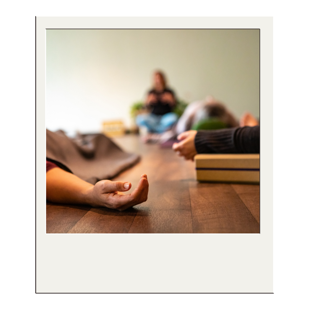 Close-up of a person’s hand in a yoga or meditation pose on a wooden table, with other people in the background participating in a meditation or yoga class.