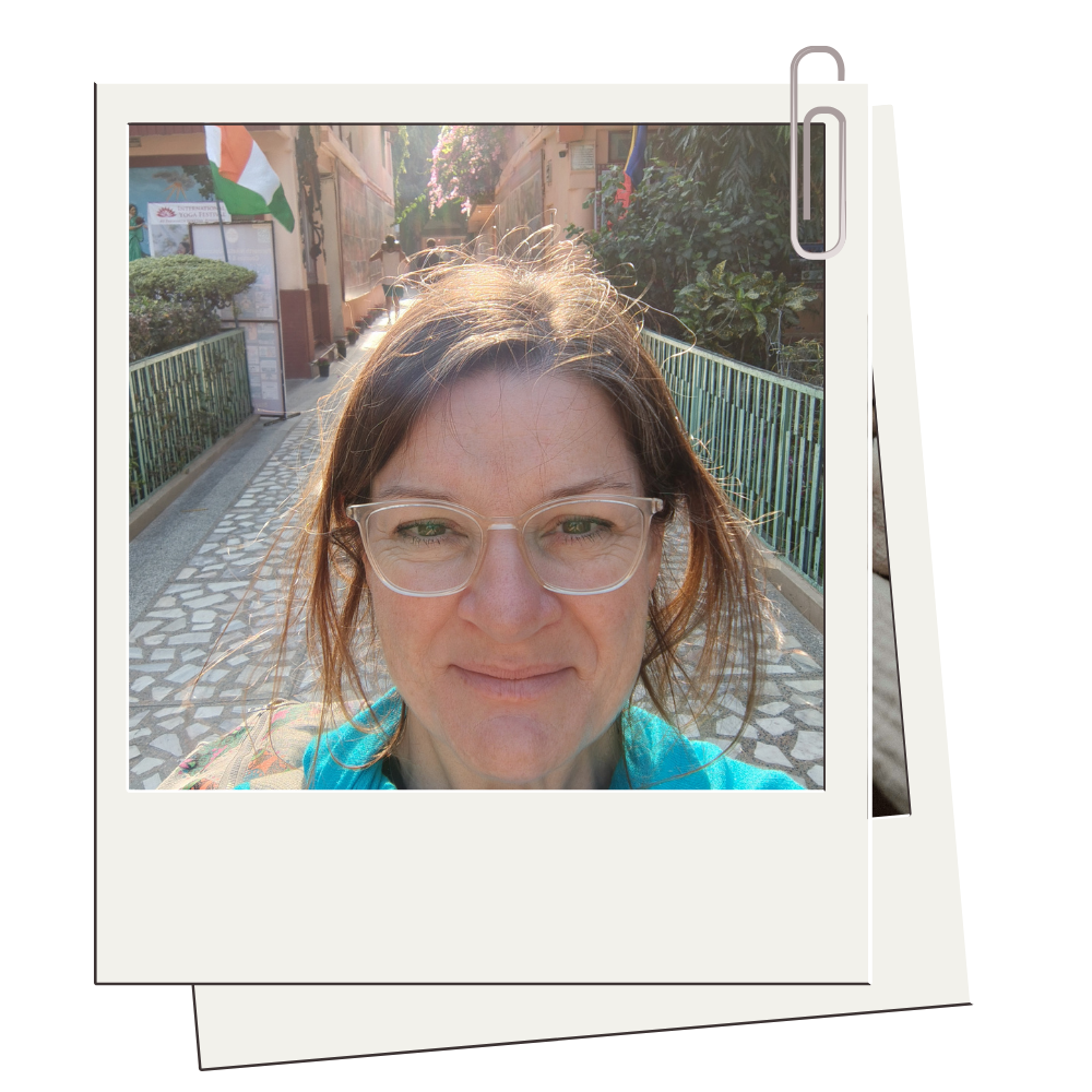 A woman with glasses smiling outdoors on a sunny day, standing on a patterned sidewalk with greenery and flags in the background.