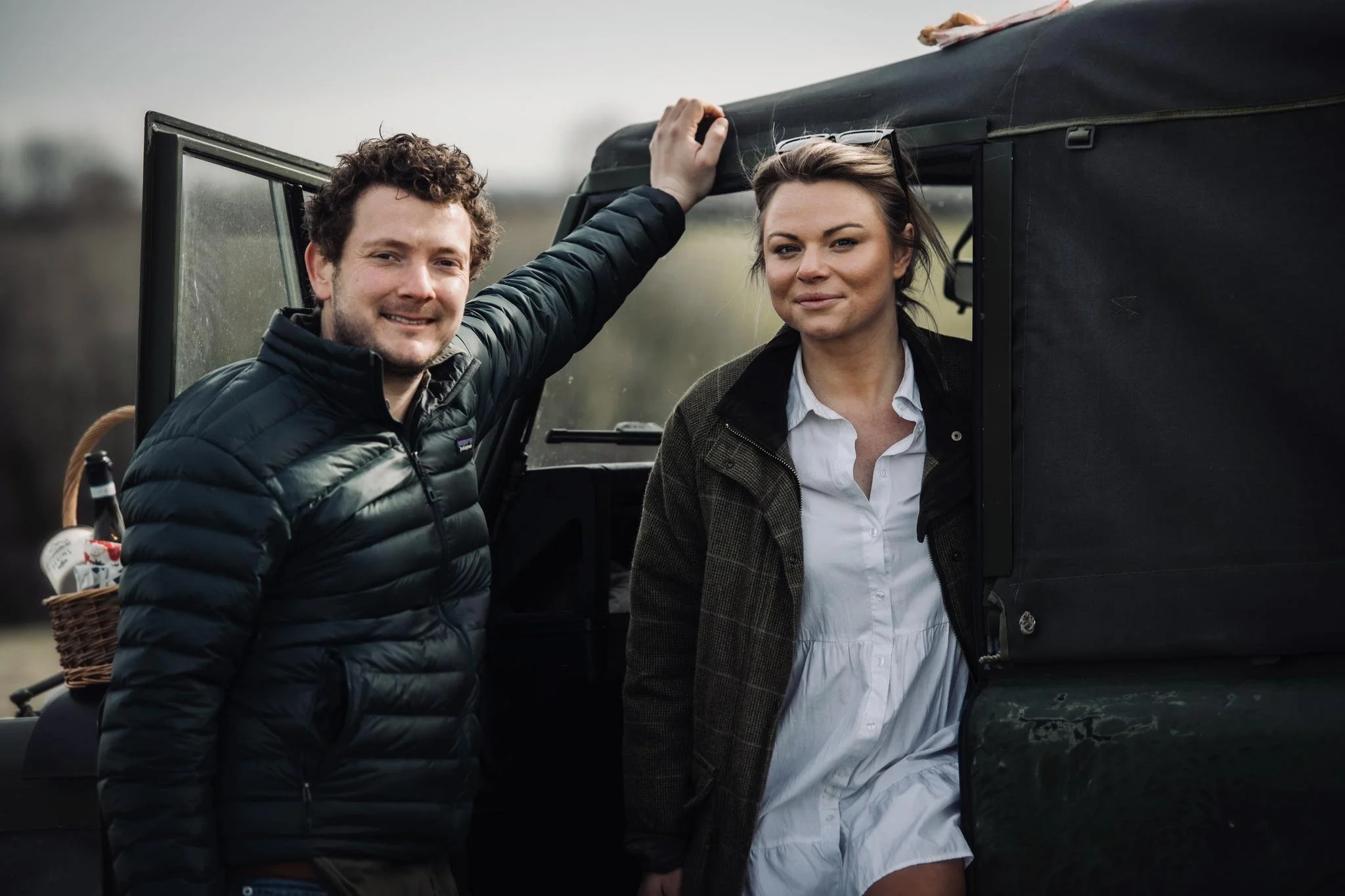 A man in a black puffer jacket and a woman in a white dress and jacket standing outside near a vintage green jeep or truck on a cloudy day.