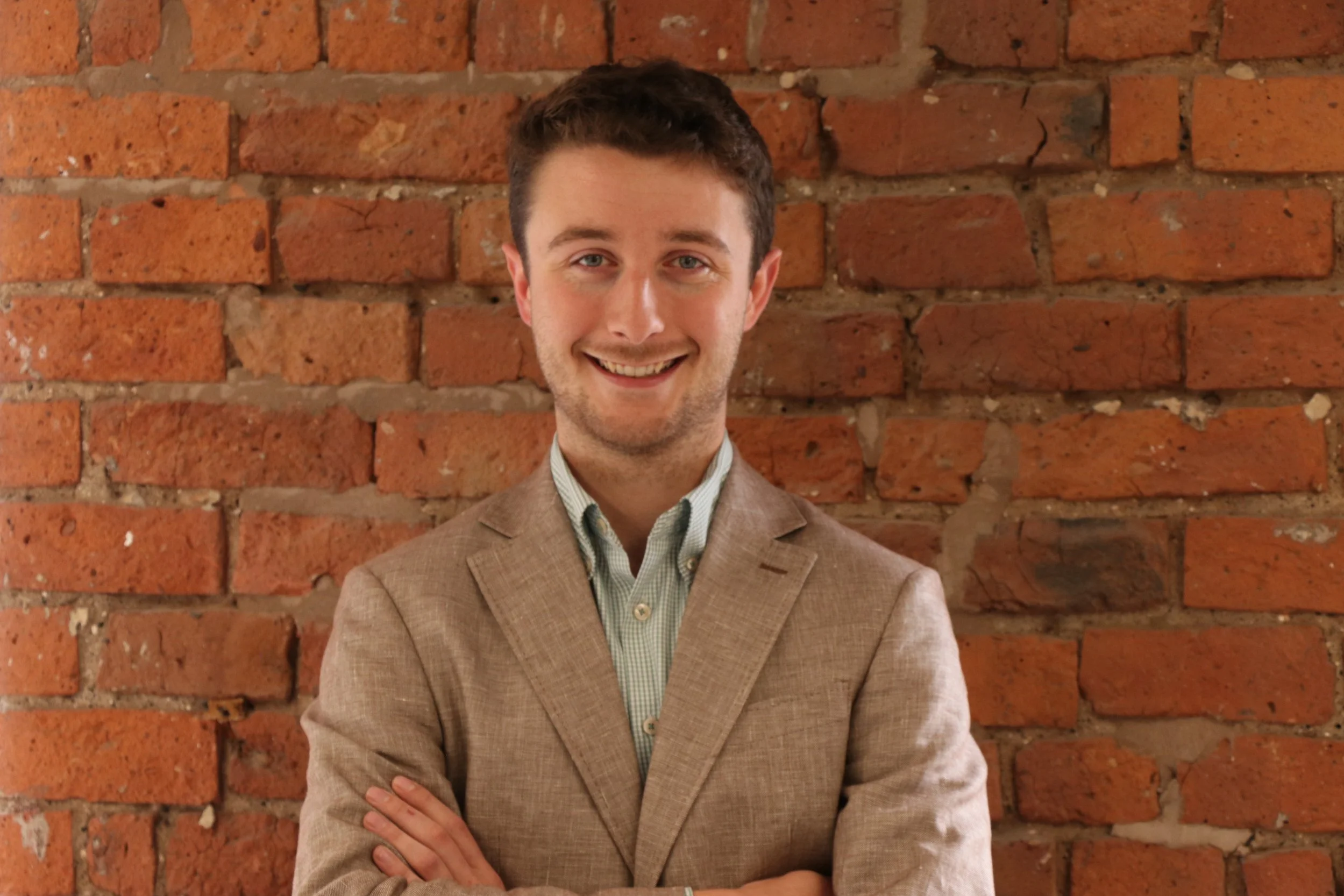 A young man with short brown hair and blue eyes, wearing a light brown suit and a checkered shirt, smiling with arms crossed, standing in front of a red brick wall.