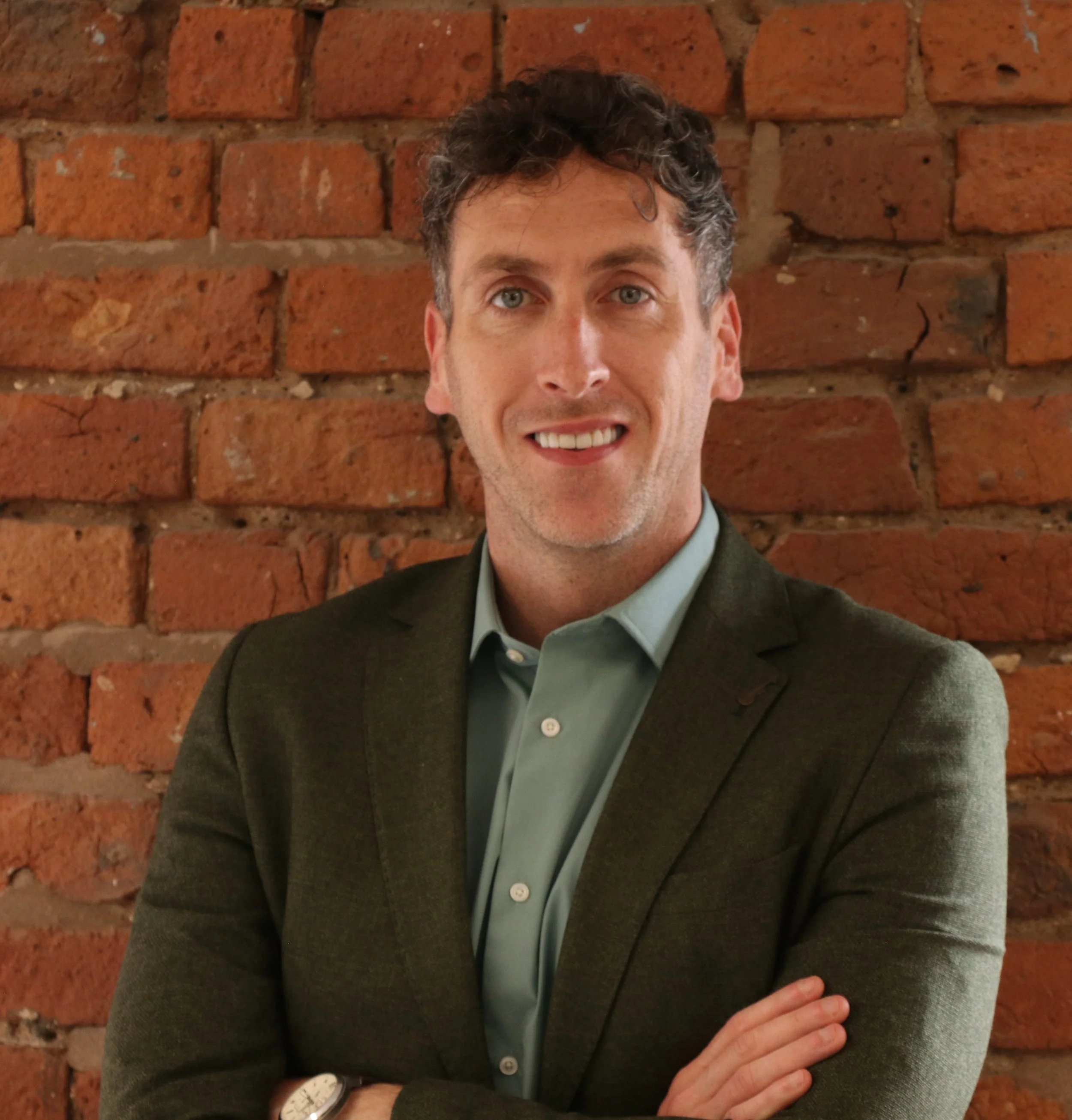 Close-up of a man smiling, wearing a suit and shirt, with crossed arms in front of a brick wall.