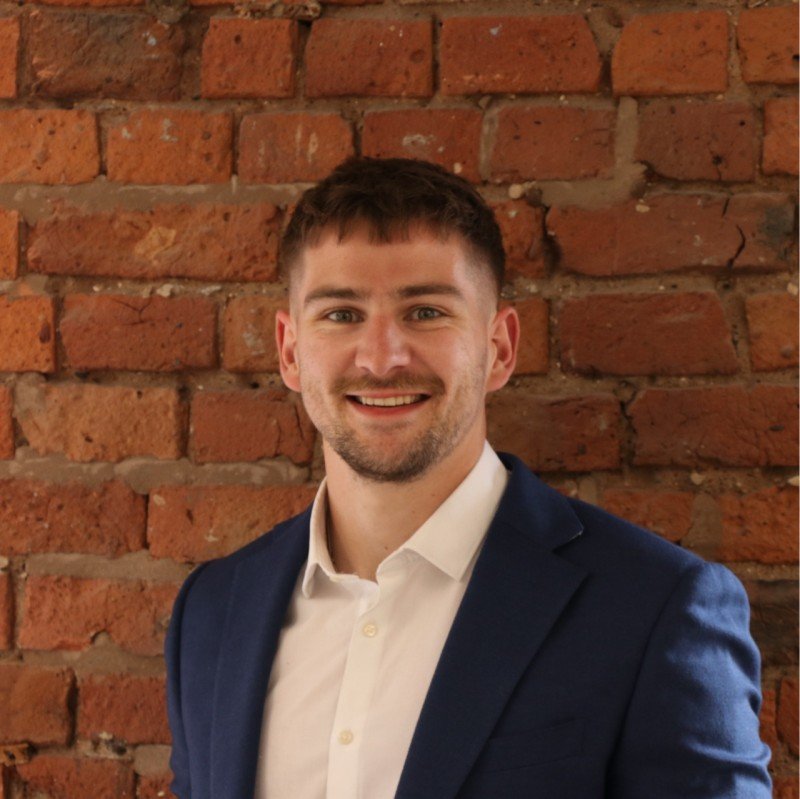 Young man in a white shirt and blue blazer smiling in front of a brick wall.