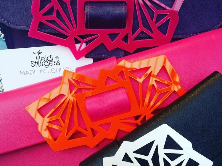 Close-up of colorful handbags with decorative buckles and tags from Heidi Sturgess, featuring pink, orange, black, and white designs.