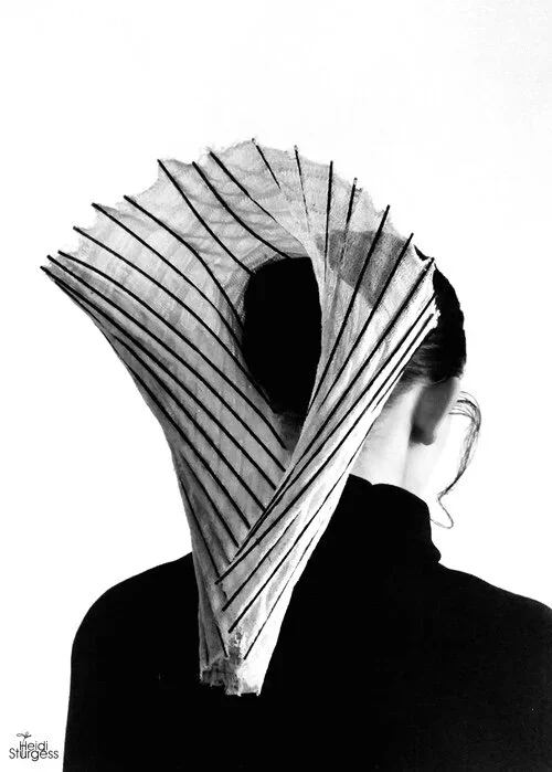 A person with their back to the camera, wearing a black shirt, has an abstract black and white paper sculpture over their head, creating a surreal art effect.