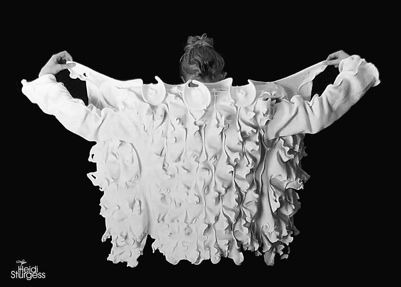 A woman with curly hair facing away, holding a large, textured, ruffled white fabric over her shoulders against a black background, with her arms extended to the sides.