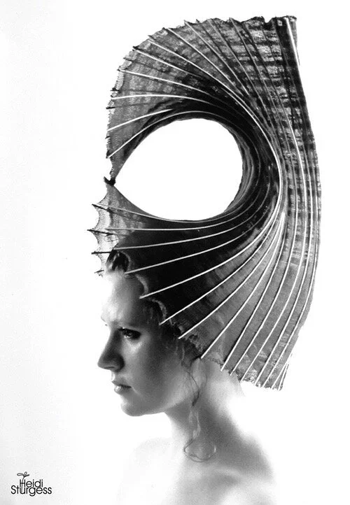 A black and white artistic photograph of a woman with her head turned to the side, wearing a large, semi-transparent, textured headpiece that has a circular opening and radiating lines.