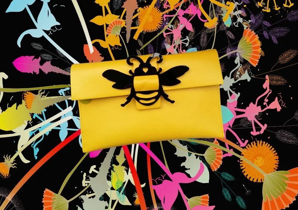 A yellow clutch purse with a black bee embellishment on the flap, placed on a vibrant floral-patterned surface with pink, orange, red, blue, and purple flowers and leaves.