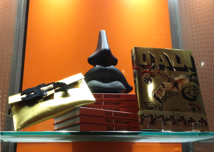Display shelf with a black abstract sculpture, gold clutch purse, red books titled "Dali/Duchamp," and a decorative large card or book with the word "Dali" and various illustrations against an orange background.