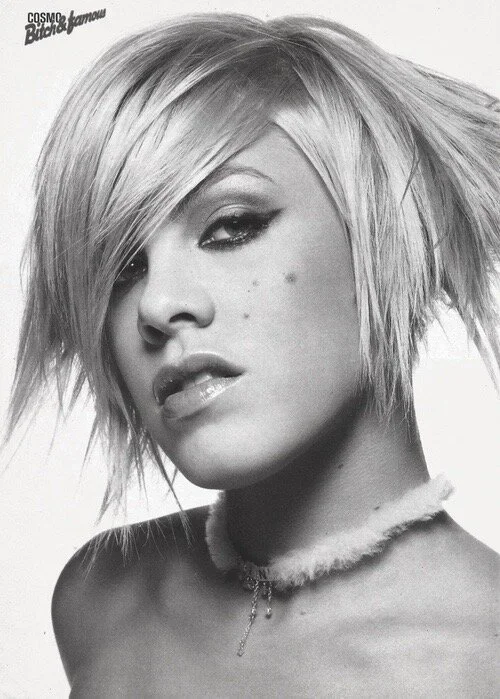 Black and white photo of a woman with messy, layered short blonde hair and a choker necklace, looking at the camera with a confident expression.