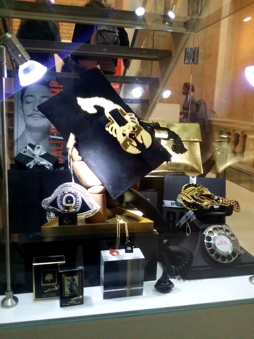 Display case with fashionable jewelry and accessories including a gold clutch purse, a silver eye-shaped ring, a gold chain, a gold bracelet, and a black rotary telephone, with a poster of a woman's face and a sign in the background.