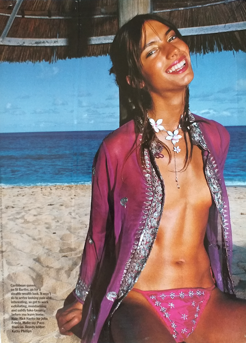 Woman smiling on a beach wearing a pink bikini and a sheer purple cover-up, with a floral necklace, in front of ocean waves and a thatched roof.