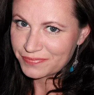 Close-up of a woman with blue eyes and dark hair, wearing earrings, smiling slightly.