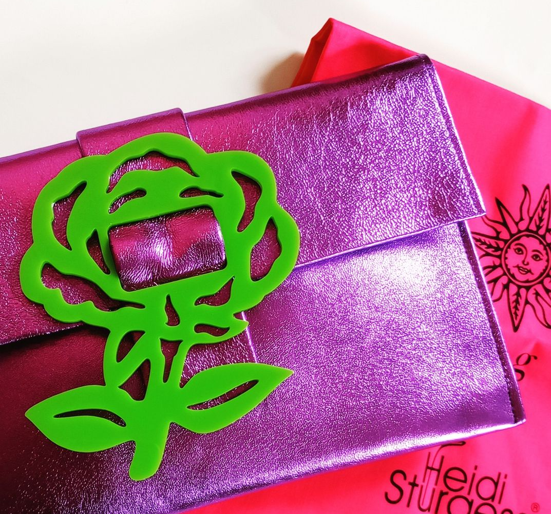 A shiny pink clutch purse with a bright green decorative flower clip attached to it, placed on a pink fabric with "Heidi Sturgeon" written on it.