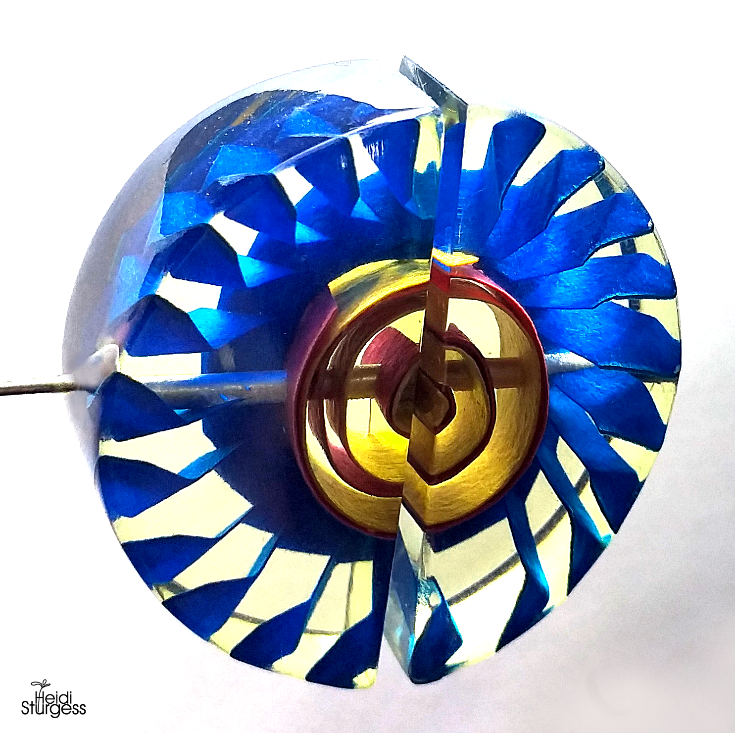 A colorful pinwheel made of glass, with blue, yellow, red, and clear sections, mounted on a metal stick against a white background.
