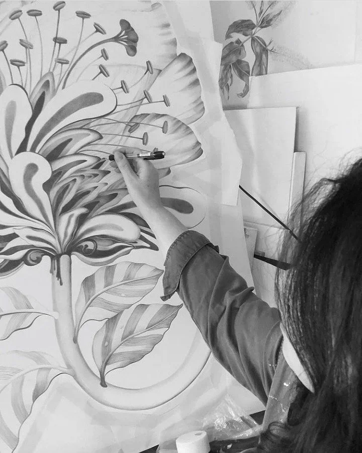 Person painting a detailed black and white botanical illustration, focusing on large leaves and flower stamens, with art supplies and papers around.