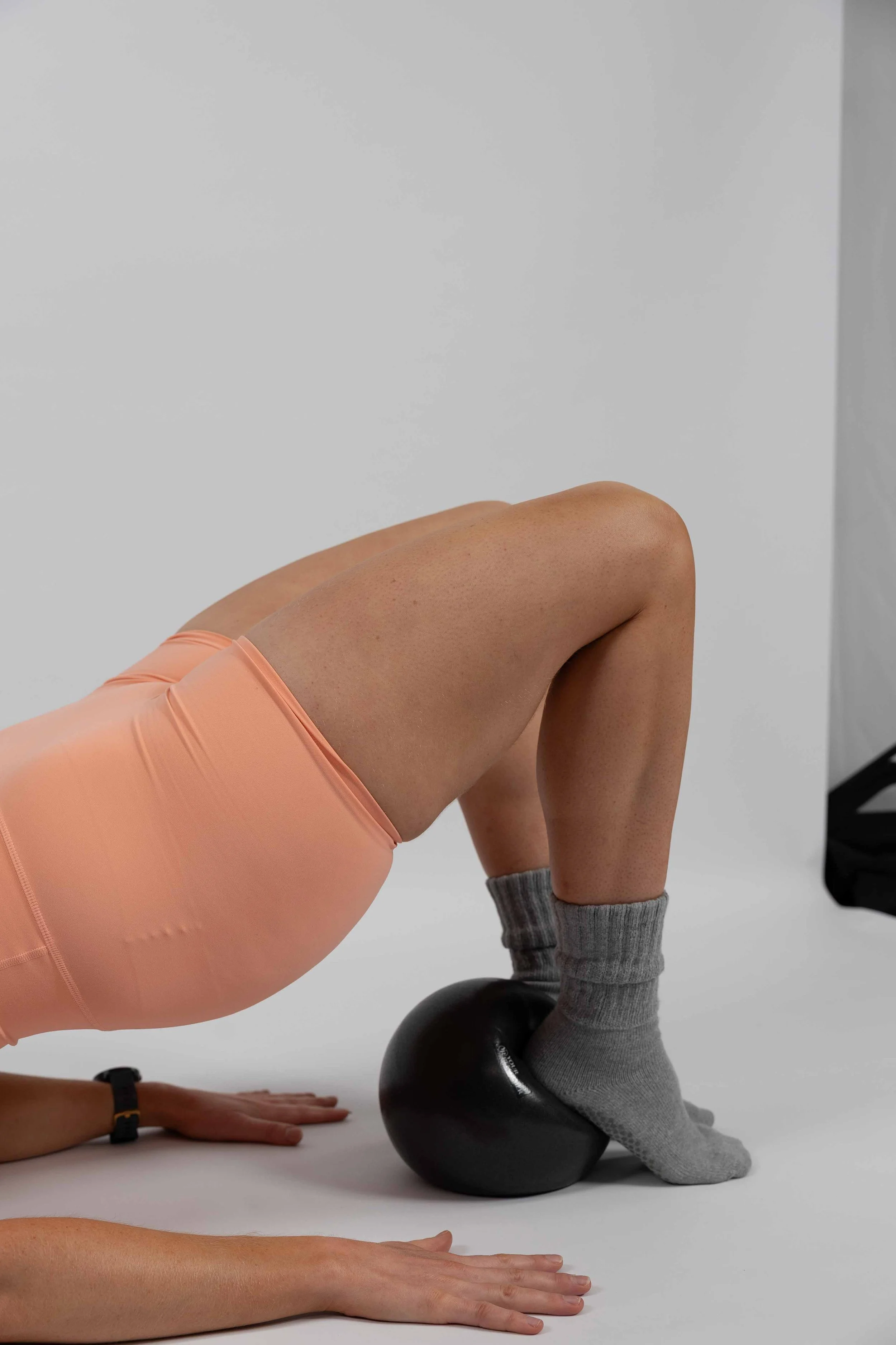 Person performing a yoga pose using a black exercise ball, with one hand on the floor and wearing gray socks and peach athletic shorts.