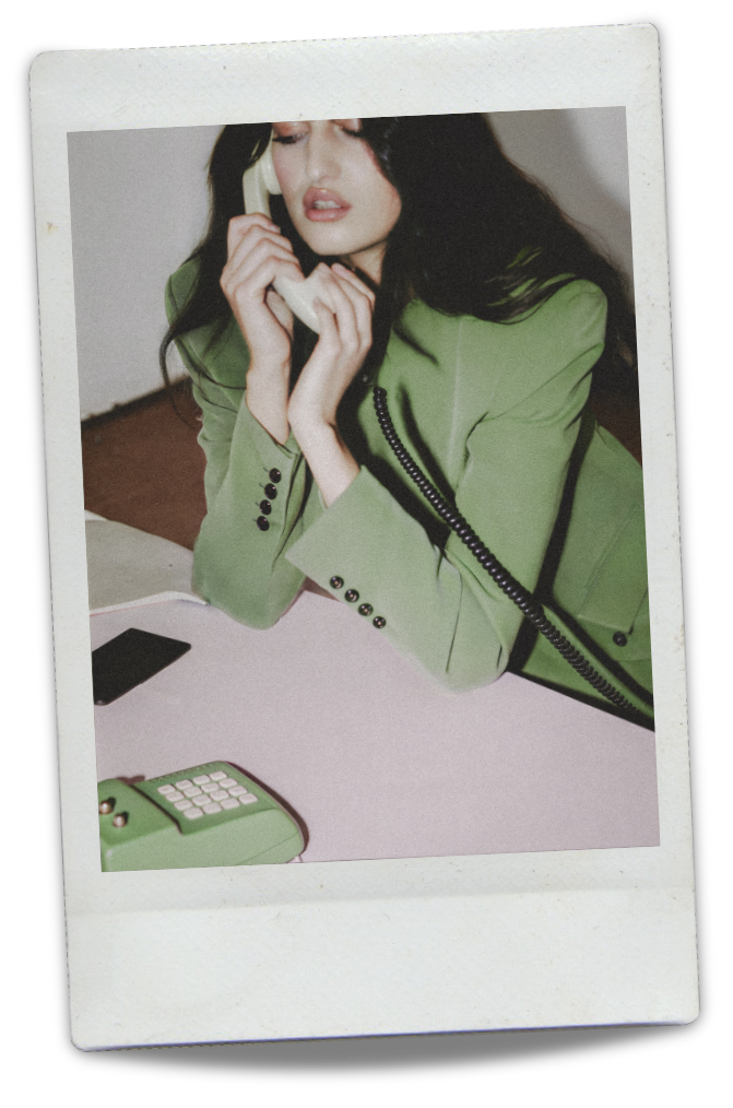 A woman with dark hair in a green blazer talking on a beige landline phone at a pink desk with a matching green rotary phone and a black object.