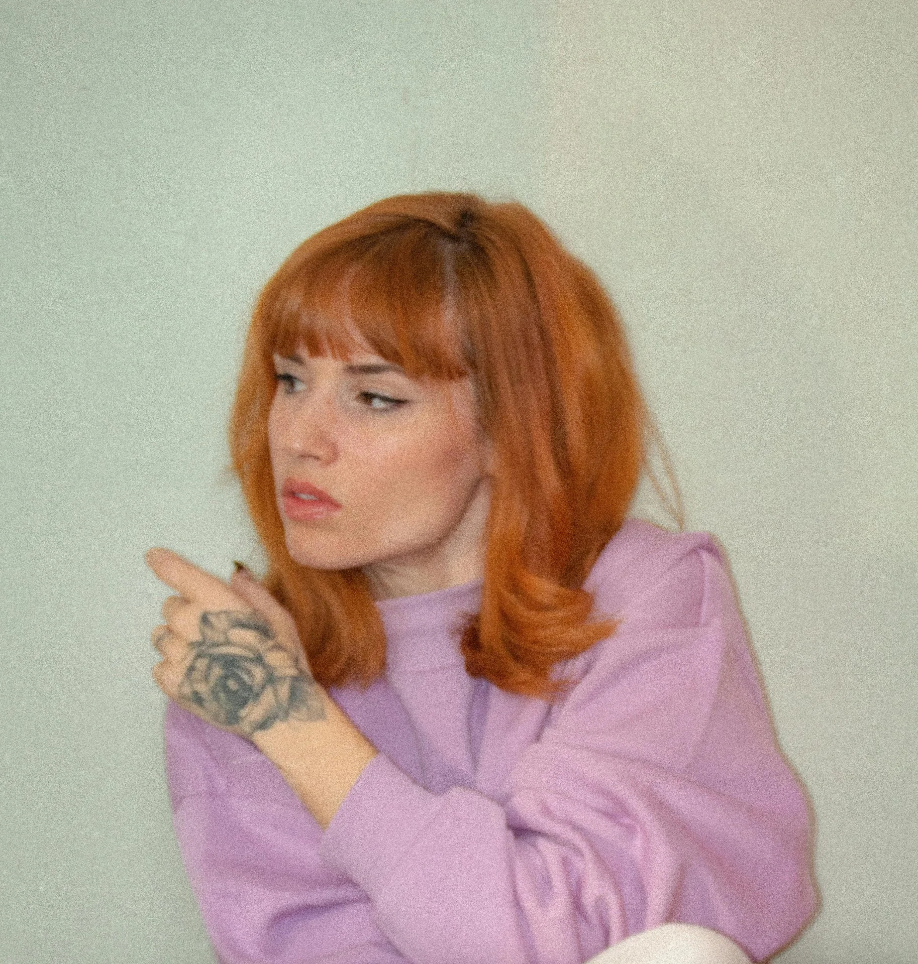 A woman with red hair and a tattoo on her arm, pointing with her finger, wearing a purple shirt, against a plain light-colored background.