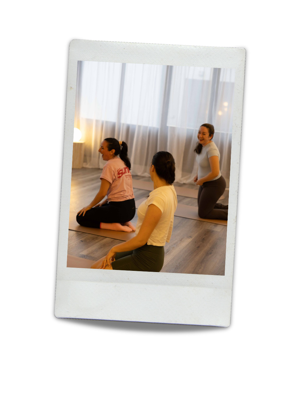 Three women participating in a yoga class in a softly lit room with white curtains, sitting or kneeling on yoga mats and smiling.
