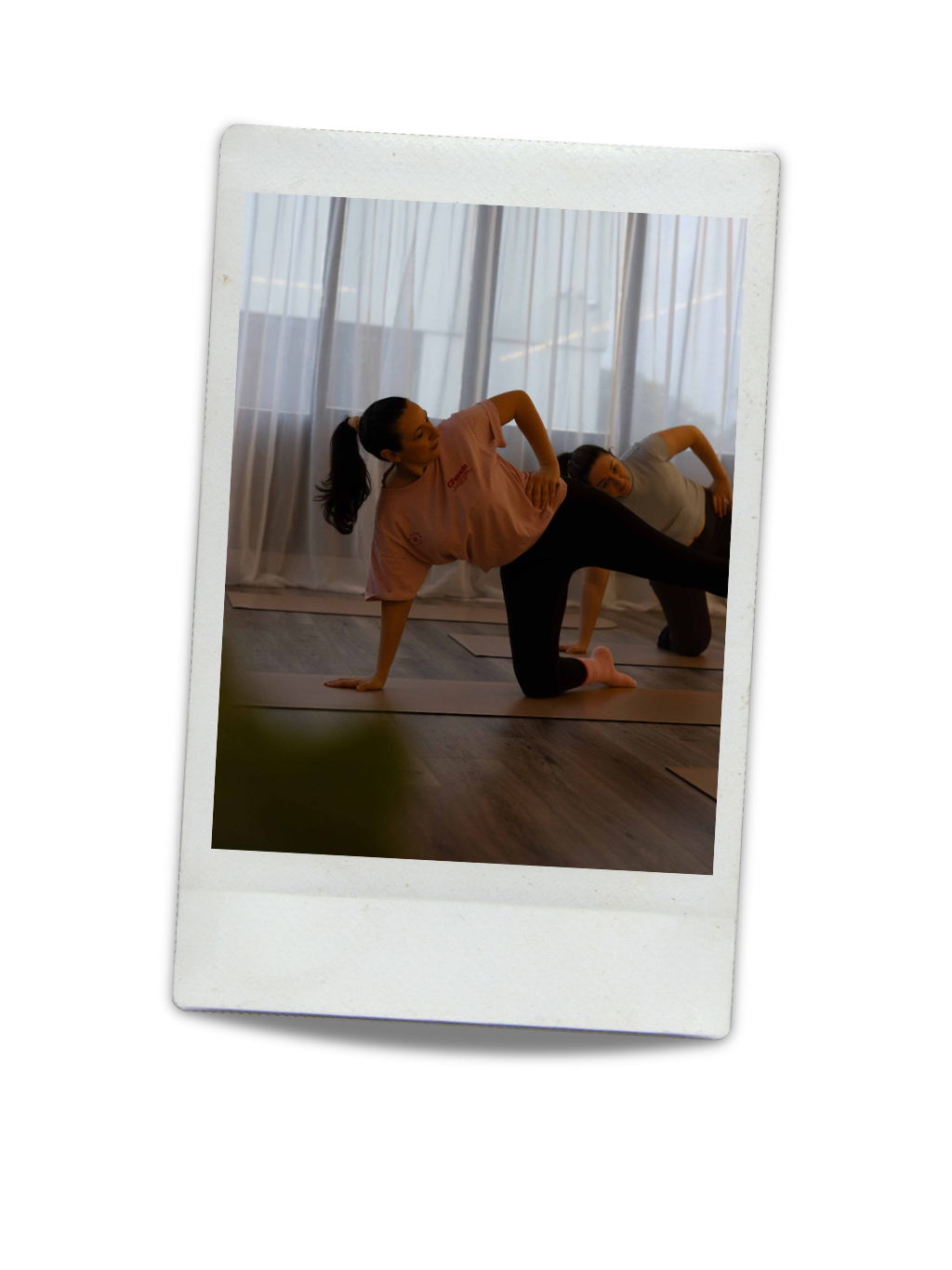 Two women practicing yoga in a studio, one in a side plank pose, with curtains and large windows in the background.