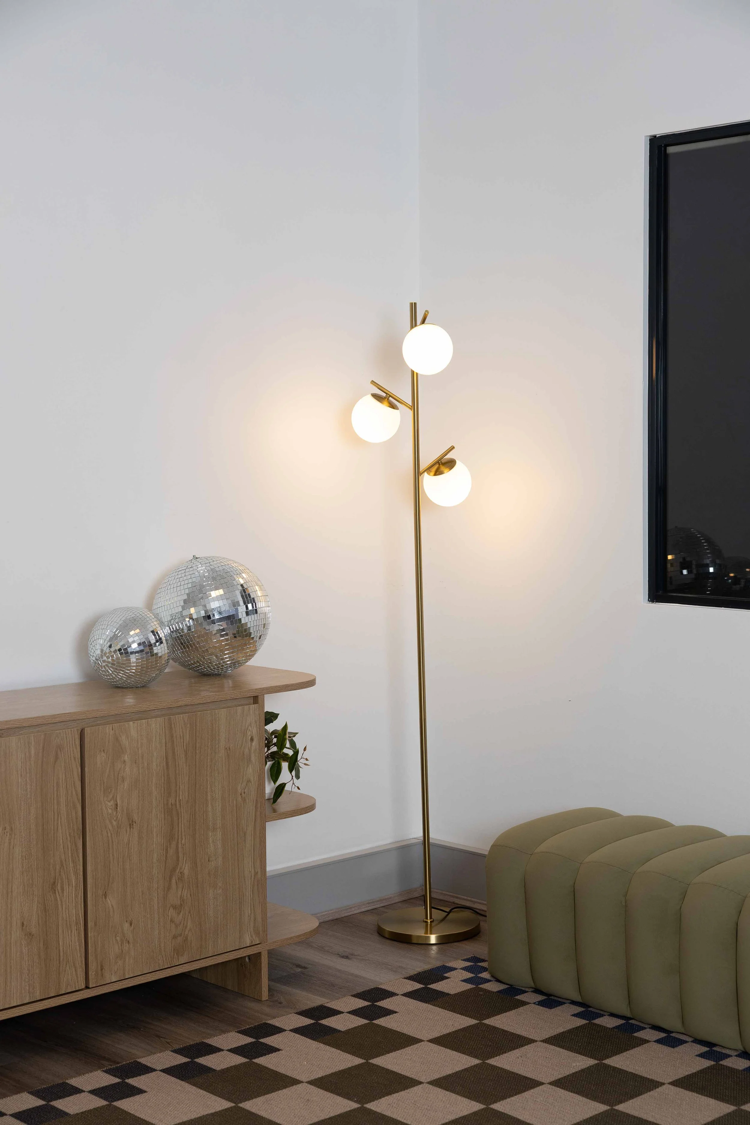 A modern living room corner with a wooden cabinet, three disco ball decorations on top, a tall gold floor lamp with three spherical lights, a light green upholstered ottoman, a black window frame, and a geometric patterned rug with shades of brown, beige, and black.
