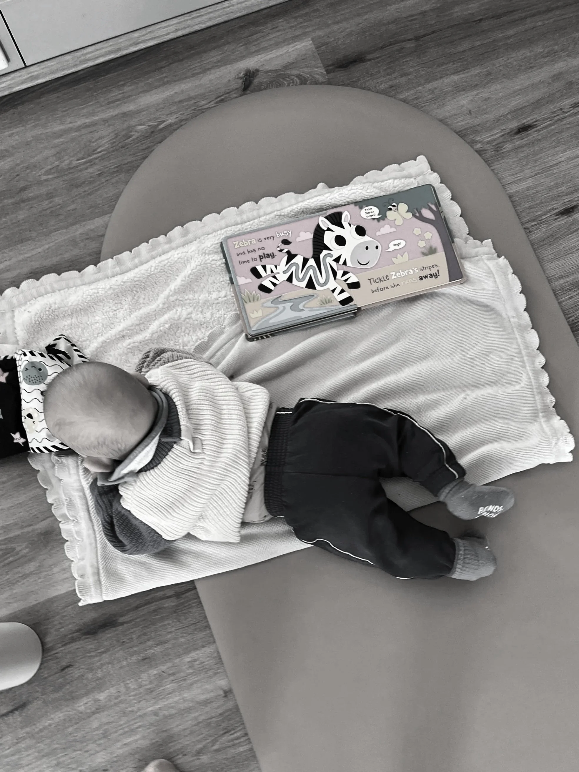 A baby lying on a bed with a white, ruffled blanket, wearing a striped shirt and dark pants, while a children's book about a zebra is open on the bed.