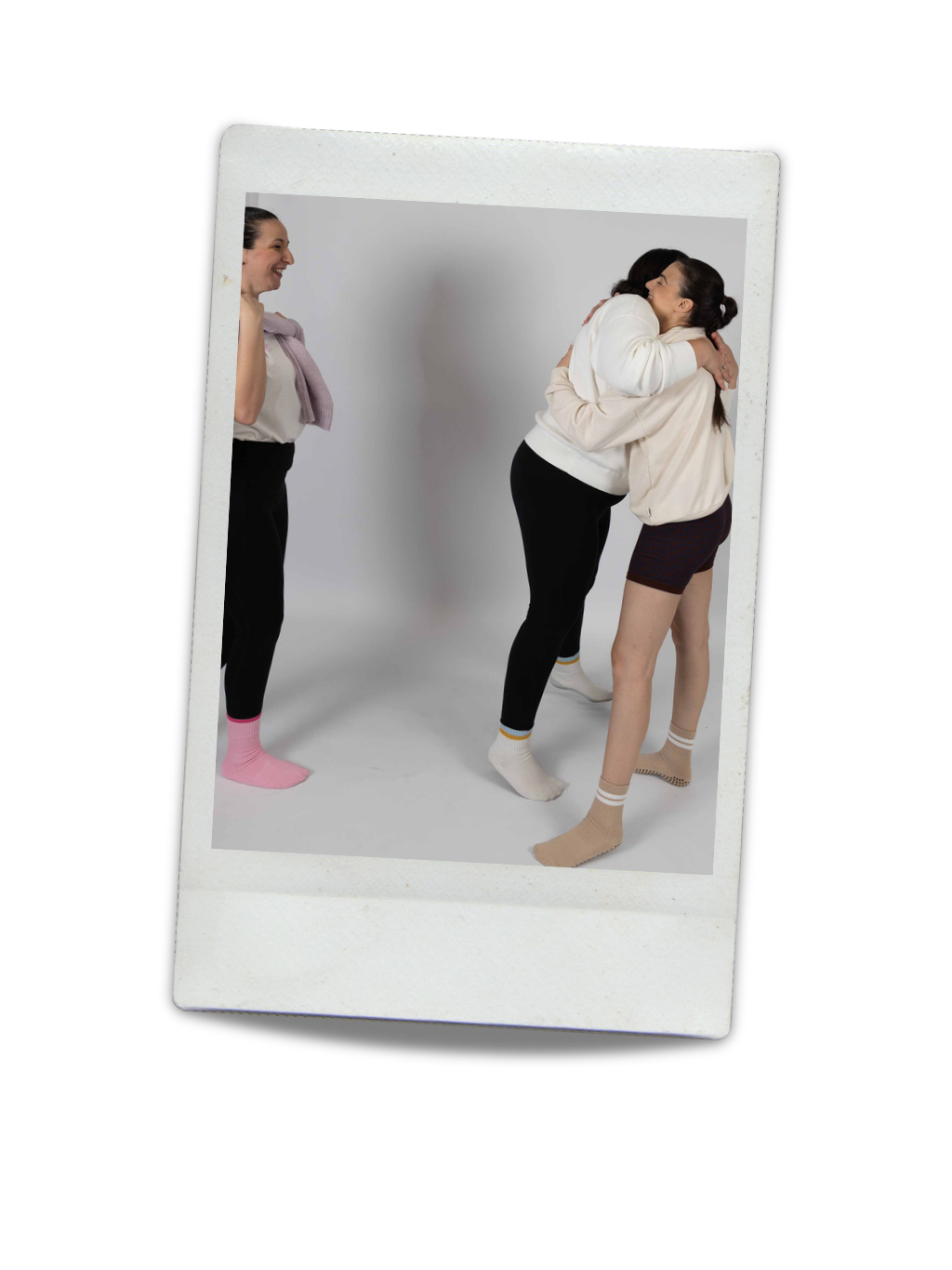 Three women in socks sharing a warm embrace and smiling, with one woman standing apart looking on in a studio with a plain background.