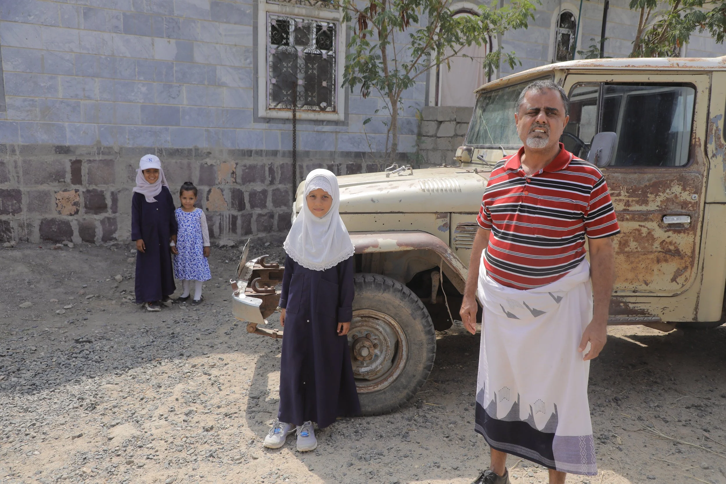 A village leader, with Ilham, Iman and Amlak, who was not two years old when her sisters encountered the mines, daughters of Radwan Mohammed, outside their home in Al Jadanah village, Shakhab, Al Dhale. An abandoned military vehicle - a remnant of th
