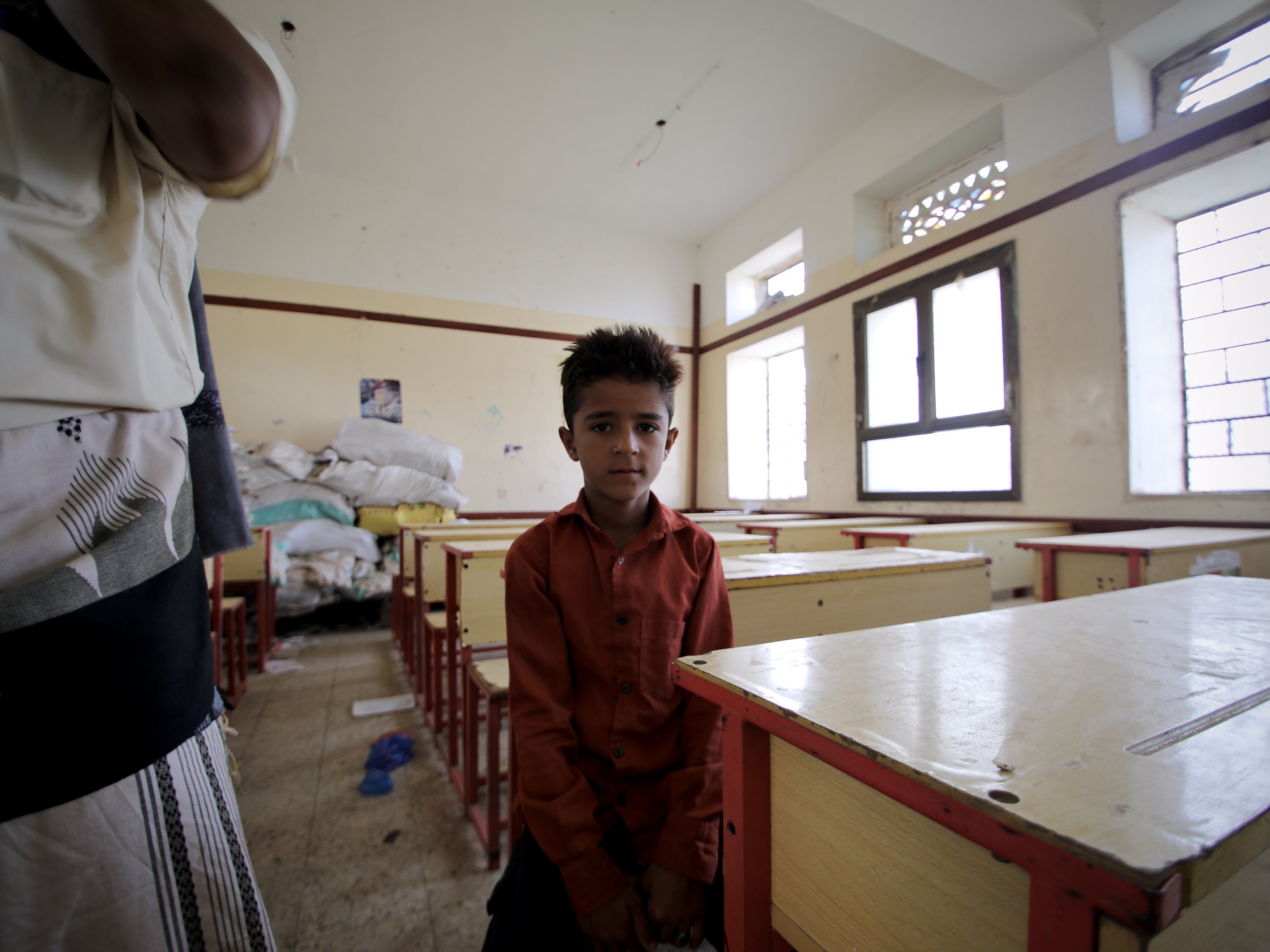 Marwan Al Barakani, 10, sits inside a classroom at Al Shaab School in Jabal Habashi district, Taiz governorate, Yemen, while his father Ahmad Mohamad Al Barakani, 33, stands nearby. After part of the school was destroyed during the conflict, around 3