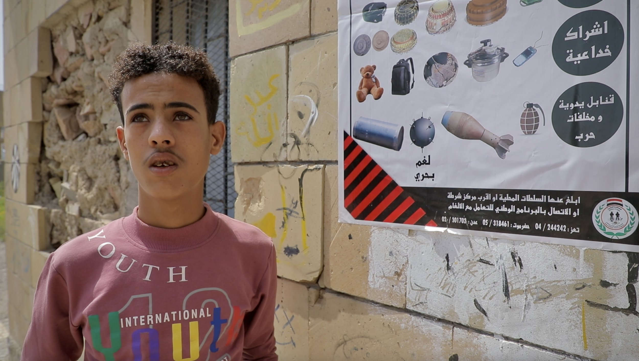 A child stands beside a warning poster illustrating explosive threats during an Explosive Ordnance Risk Education session in western Taiz, Yemen. Educators warn that landmines and improvised explosive devices may be concealed inside everyday objects,