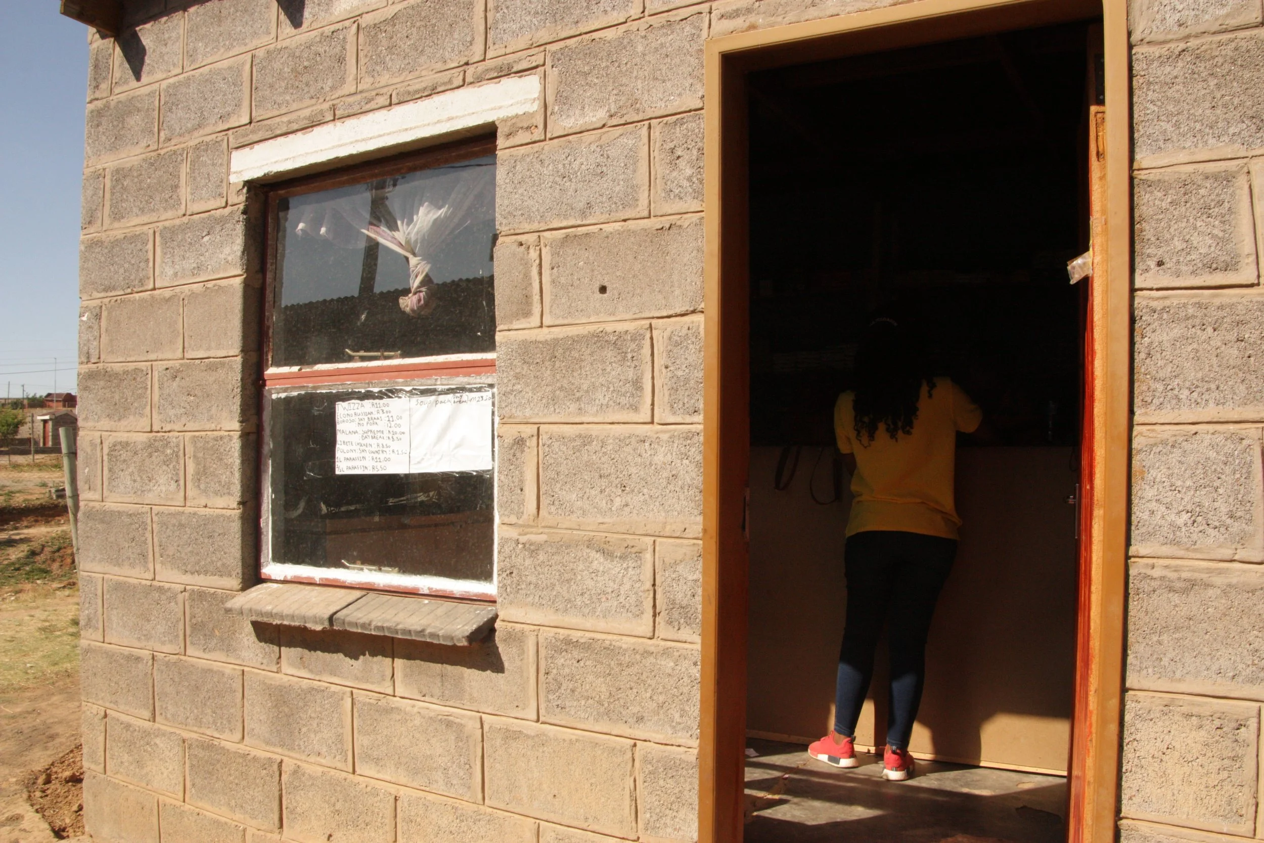 A customer stands inside the doorway of Paballo's one-room shopin Ha Magele, Leribe District, Lesotho. The business serves neighbours and schoolchildren in the area, selling essentials in small quantities to households managing rising costs and unsta